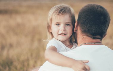 Küçük kız babasının boynuna sarılıyor ve gülümsüyor.