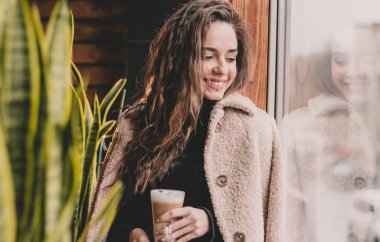 Uzun saçlı, manken görünümlü bir kadın elinde bir kupa kahveyle restoranda oturuyor ve gülümsüyor.