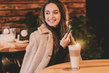 Uzun saçlı, model görünümlü bir kadın restoranda bir masada oturuyor ve elinde bir telefonla gülümsüyor.
