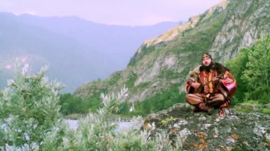 Ulusal giysili ve sırtında kurt postu olan bir adam nehrin yanındaki bir kayanın üzerinde bağdaş kurup Altai Dağları 'nın arka planına bağlı bir müzik aleti topshur çalıyor. Yavaş çekim