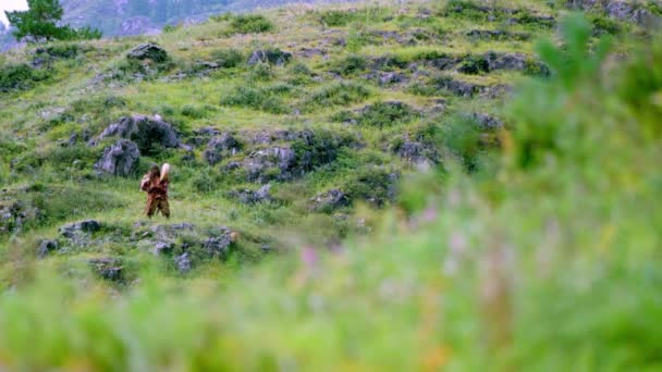 長い髪をした民族衣装を着たアルタイ族のシャーマンの役の男が顔のダンスを舞い タンバリンを打ち 腕を振っている 岩や緑の芝生の間で山の上で夏の日に撮影 — ストック動画