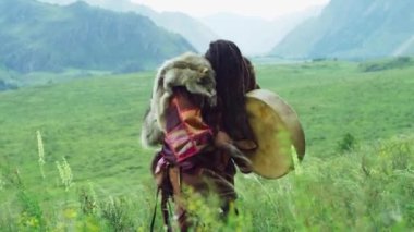 Ulusal giysili Altai şaman rolündeki adam ve omuzlarında uzun saçlarıyla dans eden bir kurt derisi, tef 'i yener. Yaz günü dağların arka planında çekim yapmak