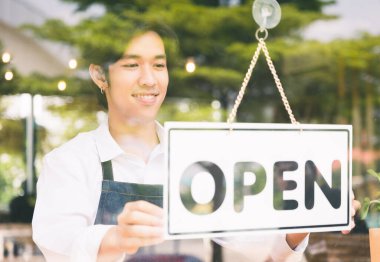Genç Asyalı adam müşteriyi karşılamak ve sabah kahve dükkanı açmak için açık etiketi çevir. Asyalı erkek barista aynadaki yansıma ile kapının önündeki kahve kafesini açıyor..