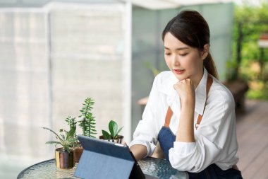 Ev bitkilerinde çalışan Asyalı genç ve tatlı bir kadın. Asyalı kadın bahçıvan, seradaki ağaçların büyümesini kaydetmek ve yakalamak için tablet kullanıyor. Ev konseptinden küçük işletme çalışmaları başlat.