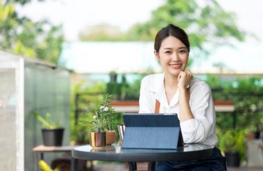 Ev bitkilerinde çalışan Asyalı genç ve tatlı bir kadın. Asyalı kadın bahçıvan, seradaki ağaçların büyümesini kaydetmek ve yakalamak için tablet kullanıyor. Ev konseptinden küçük işletme çalışmaları başlat.