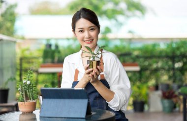 Ev bitkilerinde çalışan Asyalı genç ve tatlı bir kadın. Asyalı kadın bahçıvan, seradaki ağaçların büyümesini kaydetmek ve yakalamak için tablet kullanıyor. Ev konseptinden küçük işletme çalışmaları başlat.
