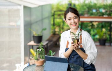 Ev bitkilerinde çalışan Asyalı genç ve tatlı bir kadın. Asyalı kadın bahçıvan, seradaki ağaçların büyümesini kaydetmek ve yakalamak için tablet kullanıyor. Ev konseptinden küçük işletme çalışmaları başlat.
