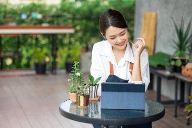 Ev bitkilerinde çalışan Asyalı genç ve tatlı bir kadın. Asyalı kadın bahçıvan, seradaki ağaçların büyümesini kaydetmek ve yakalamak için tablet kullanıyor. Ev konseptinden küçük işletme çalışmaları başlat.