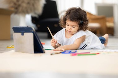 Tabletle öğrenilen büyük kağıtta suluboya boyayla resim yapan tatlı bir kız. Oturma odasında yerde oturuyor. Evde mutlu çocuk anları. Anaokulunda sanat etkinliği.