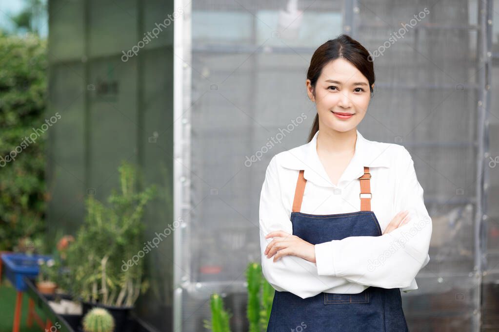 Young cute asian woman in uniform working in house plant standing with arm crossed. Asian female taking care of the tree in greenhouse. Startup small business work from home concept.