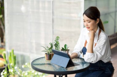 Ev bitkilerinde çalışan Asyalı genç ve tatlı bir kadın. Asyalı kadın bahçıvan, seradaki ağaçların büyümesini kaydetmek ve yakalamak için tablet kullanıyor. Ev konseptinden küçük işletme çalışmaları başlat.