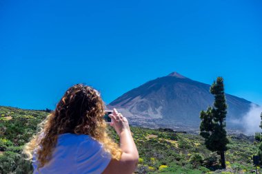 Young tourist woman observing a flower in Del Teide National Park. In the background is the Teide. This volcano is located in Tenerife, Canary Islands. clipart