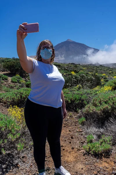 Young tourist woman with mask taking a selfie. In the background you can see Mount Teide, with its 3,178 meters high. This volcano is located in Tenerife, Canary Islands.