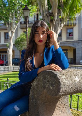 Bir portre için poz veren genç bir kadın. Amerikan ceketi, kot pantolon ve topuklu ayakkabı giyiyor. La Laguna, Tenerife 'de bir bankta oturuyor..