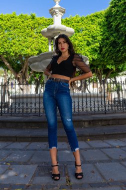 Bir portre için poz veren genç bir kadın. Siyah bir gömlek, kot pantolon ve topuklu ayakkabı giyiyor. Arka planda La Laguna, Tenerife 'de bir meydanda bir çeşme var..