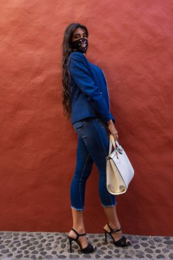 Young woman leaning against a red wall holding a purse with mask. She wears high heel shoes, jeans and an American jacket. The photo was taken in La Laguna, Tenerife.