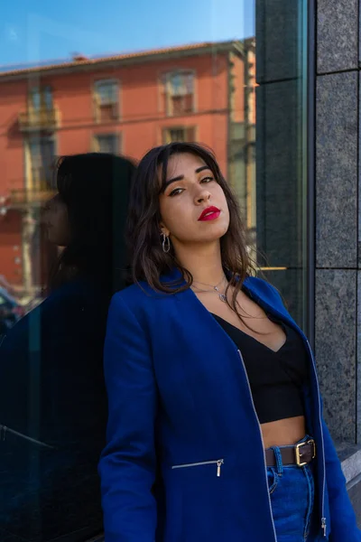 Young woman posing for a portrait in the background looks a glass. She wears an American jacket and jeans. The photo was taken in La Laguna, Tenerife.