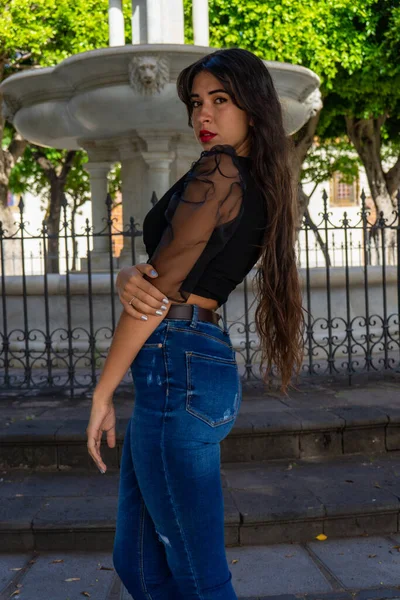 年轻女子摆出一副画像的姿势 她穿着一件黑色衬衫 牛仔裤和高跟鞋 在Tenerife的La Laguna的一个广场上有一个喷泉 — 图库照片