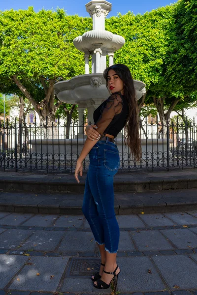 年轻女子摆出一副画像的姿势 她穿着一件黑色衬衫 牛仔裤和高跟鞋 在Tenerife的La Laguna的一个广场上有一个喷泉 — 图库照片