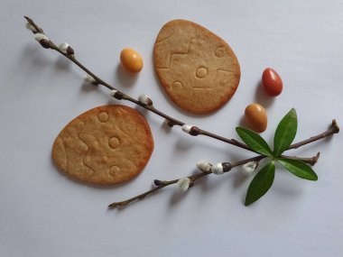 Paskalya geçmişi. Yumurta şeklinde kurabiyeler, bahar ağacı dalları, beyaz arka planda renkli yuvarlak şekerler. Paskalya yemeği, kurabiye.