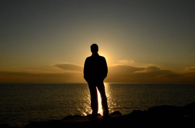 Siluet Genç adam kayalıklarda gün batımında sahilde, yalnız.