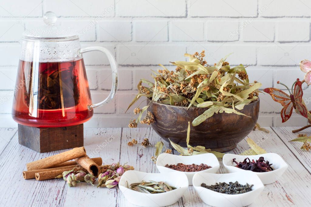A variety of herbs brewed in a glass teapot on wood in front of the white wall. Linden, pomegranate flower, green tea, grape seed, olive leaf, cinnamon sticks and dried rose in a glass cup.