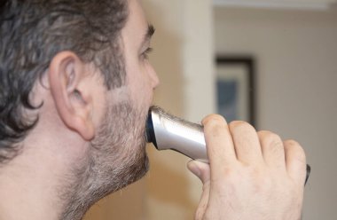 Bir adam salgında evinde sakalını şarj edilebilir makineyle tıraş ediyor.