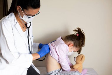 Doktoru tarafından pediatrik muayenede olan tombul küçük kız. Doktor akciğerlerini ve kalbini dinler. Koruyucu yüz maskeleri takıyorlar.
