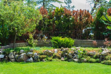 Taştan bir duvar ve demir bir çitle çevrili büyük taşlar ve bitkilerle süslenmiş bir bahçe..