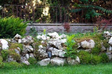 Taştan bir duvar ve demir bir çitle çevrili büyük taşlar ve bitkilerle süslenmiş bir bahçe..