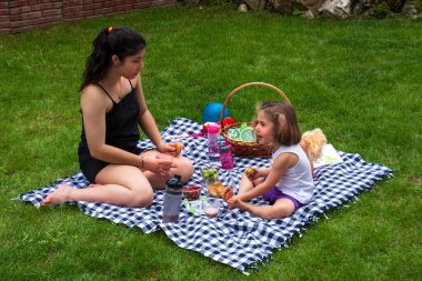 Küçük kız ve kız kardeşi oyun oynuyorlar ve bahçede piknik yapıyorlar.