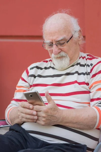 old man sitting in front of his house, dealing with his cell phone. can hardly see.