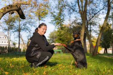 Genç bir kadın bir dobermanla yürüyüş yapıyor ve evcil bir hayvanla konuşmaktan zevk alıyor. Bulanık arkaplanı olan seçici odaklanma. Sığ alan derinliği.