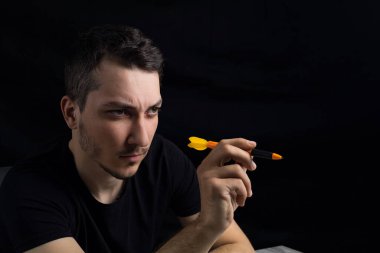 Siyah elbiseli genç yakışıklı bir adam elinde dart uçlu bir kalem tutuyor. Alçak bir anahtarın siyah arka planında. Ne pahasına olursa olsun zafer