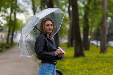 Şehir parkının bulanık arka planında şeffaf şemsiyesi olan genç bir kadın. Metin için alanı kopyala