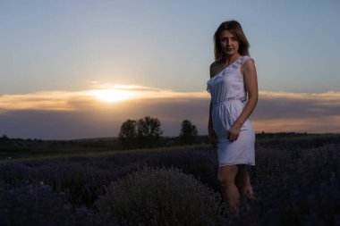Beyaz elbiseli güzel ve güzel genç bir kadın gün batımında lavanta tarlasında yürüyüş yapmaktan hoşlanıyor. Bulanık arkaplan ile seçmeli odak.