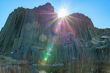 Sunlit geometric basalt columns with lens flare and clear blue sky.