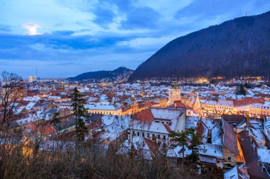 A city at night with a full moon in the sky. The city is lit up with lights and the buildings are tall. The sky is cloudy and the moon is visible in the background