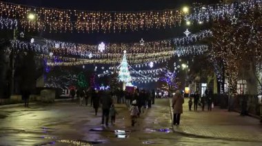 İnsanlar kış tatilinde parlak meydanlarda çelenklerle ve parlayan ağaçlarla yürüyorlar. 27 Aralık 2024 Balti Moldova.