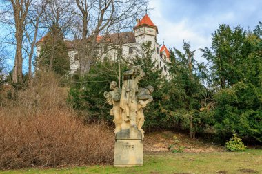 Kalenin önündeki tarihi taş heykel, yemyeşil ağaçlar ve mavi gökyüzü arasında. 17 Mart 2025, Konopiste Şatosu, Çek Cumhuriyeti.