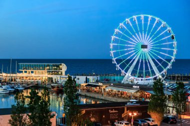 Alacakaranlıkta limanda tekneler ve modern binalarla aydınlatılmış dönme dolap. 3 Mayıs 2025 Romanya.