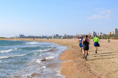 Arka planda şehir manzarasıyla güneşli sahilde koşan bir grup adam. 4 Mayıs 2025 Romanya.
