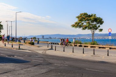 Sahil yolu boyunca insanların gezindiği güneşli sahil yürüyüşü ve manzaralı ufuk manzarası. 1 Ağustos 2025, Nessebar Bulgaristan.
