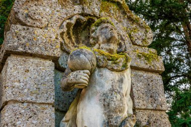 Yıpranmış taş heykel, gür bir orman ortamında küreyi tutan bir melek heykeli. 17 Mart 2025, Konopiste Şatosu, Çek Cumhuriyeti.