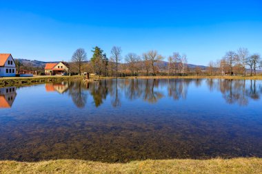 Kırsal alanda berrak mavi gökyüzü ve yansımaları olan sakin bir göl. 21 Mart 2025, Holasovice, Çek.