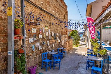 Mavi sandalyeleri ve duvar süslemeleri olan renkli kaldırım taşı sokaktaki büyüleyici açık hava kafesi. 3 Ağustos 2025, Ayvalik, Türkiye.