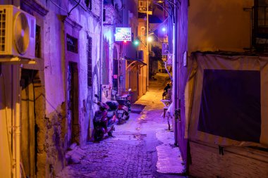 Dar kaldırım taşı caddesinde, neon ışıklı ve park etmiş scooterlı sessiz bir gece manzarası. 3 Ağustos 2025, Ayvalik, Türkiye.