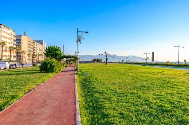 Güneşli bir patika ve deniz kenarında yeşil çimenler. Arka planda şehir manzarası var. 4 Ağustos 2025 İzmir Türkiye.