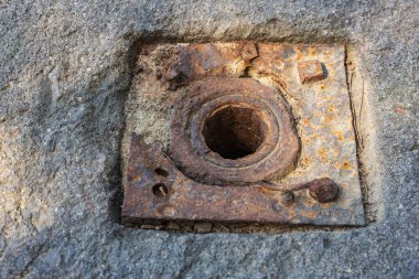 Merkezi delik ve cıvatalarla beton yüzeyde paslı metal plaka.