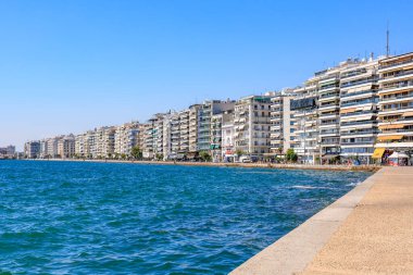 Mavi okyanusu ve binaları olan bir sahil. Binalar yüksek ve balkonları var. Sahil insanlarla dolu ve oturabilecekleri banklar var. 8 Ağustos 2024 Selanik Yunanistan
